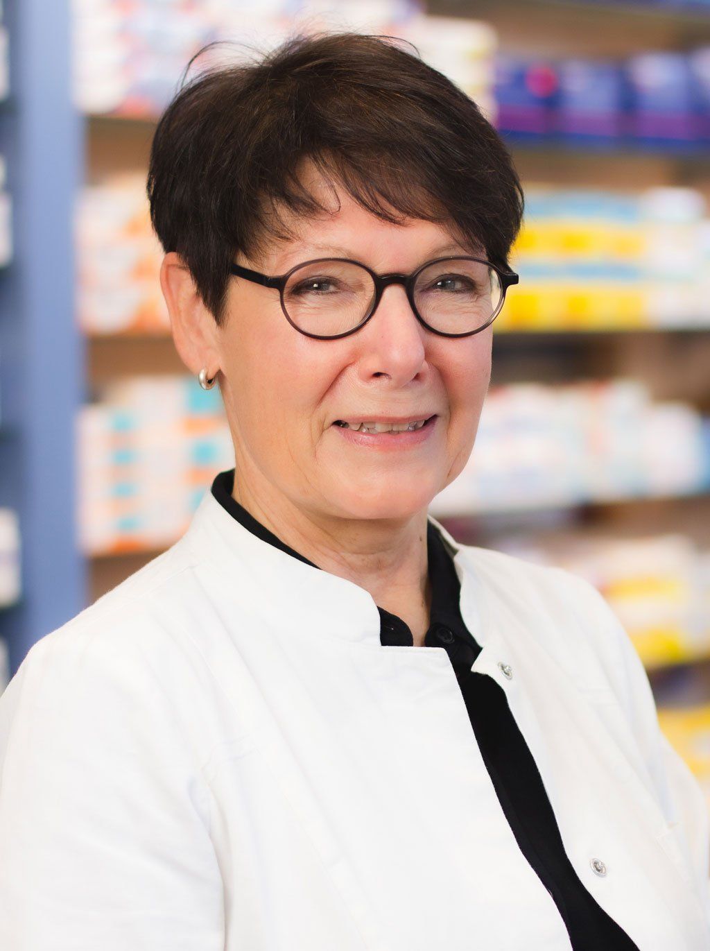 Eine Frau mit Brille und weißem Kittel steht vor einem Apothekenregal.