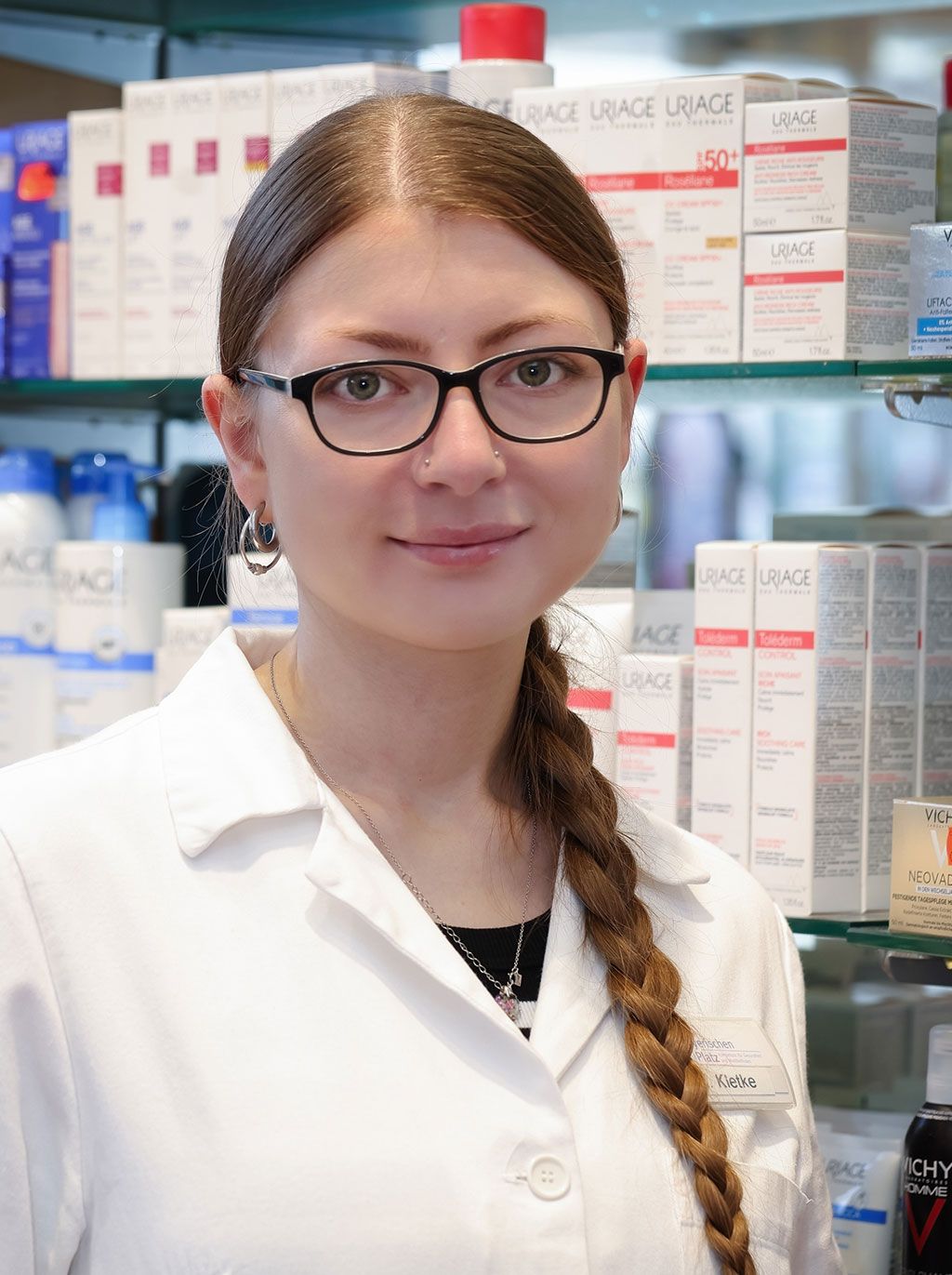 Eine Frau mit Brille und weißem Kittel steht vor einem Regal in einer Apotheke.