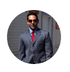 Man in a gray suit with a red tie and sunglasses standing outdoors in sunlight