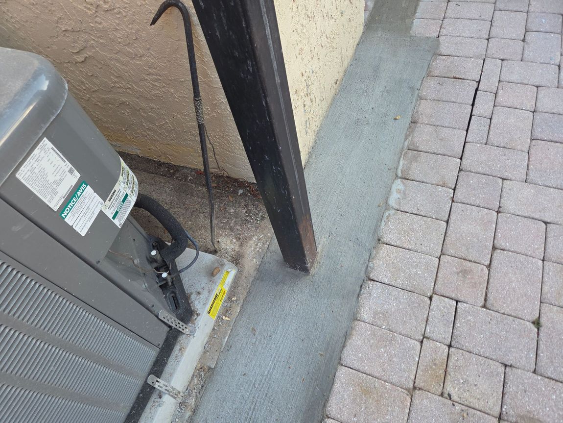 AC unit next to a wall, conduit, and paved walkway. Gray concrete area in between.