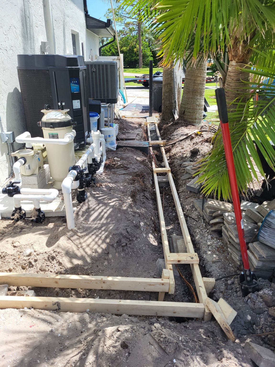 Construction site alongside a building with pool equipment. Wooden forms in a trench with a shovel and palm trees.
