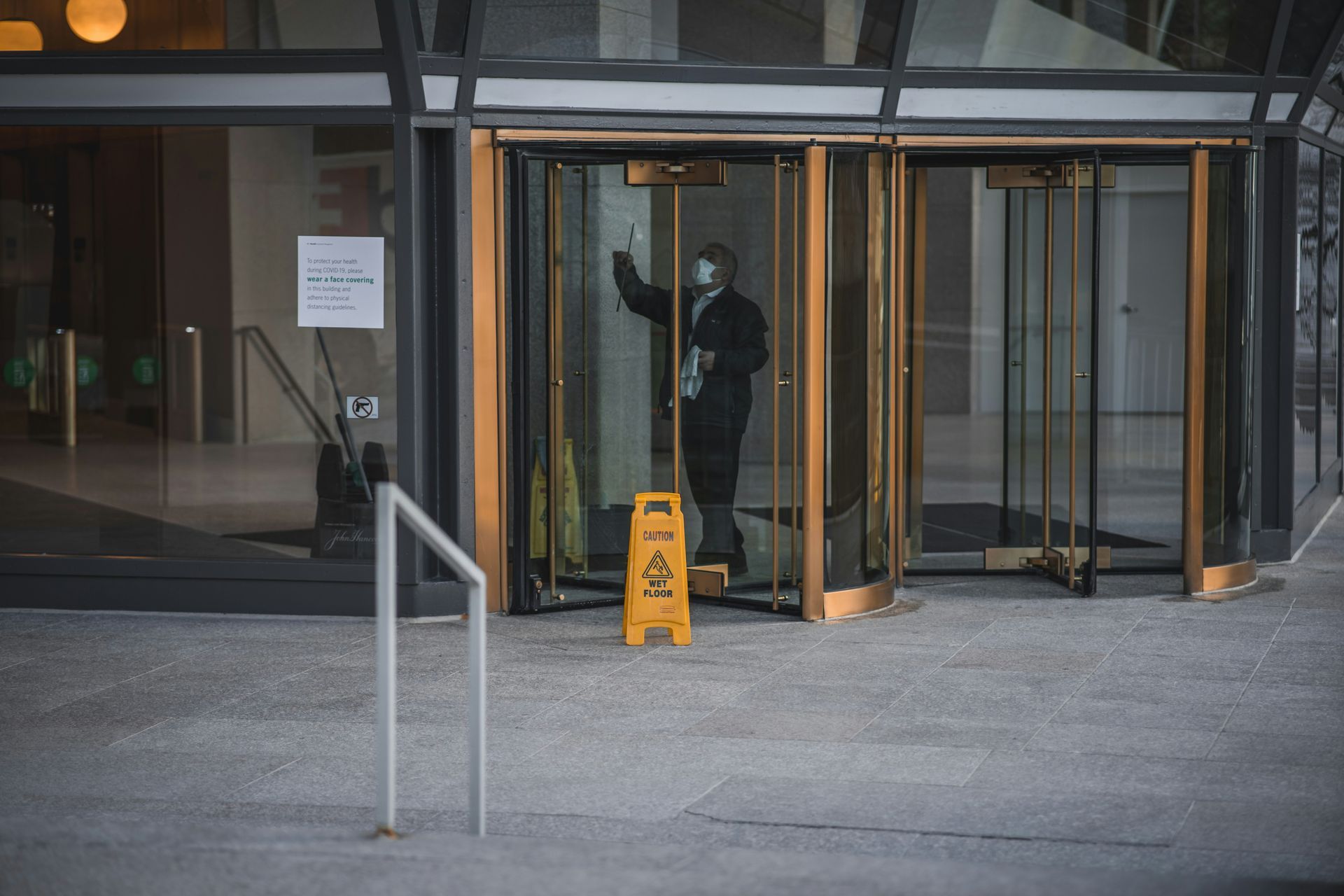 A janitor in a blue suit cleaning the glass door panels of a commercial building lobby.
