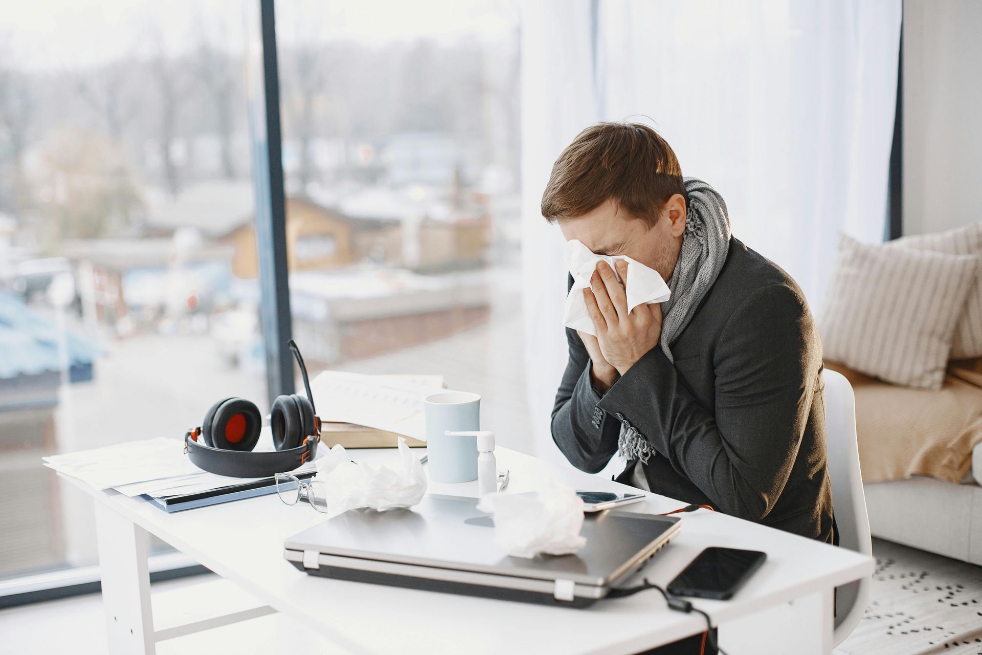 A man sitting on a desk, sniffling and sneezing with a tissue.