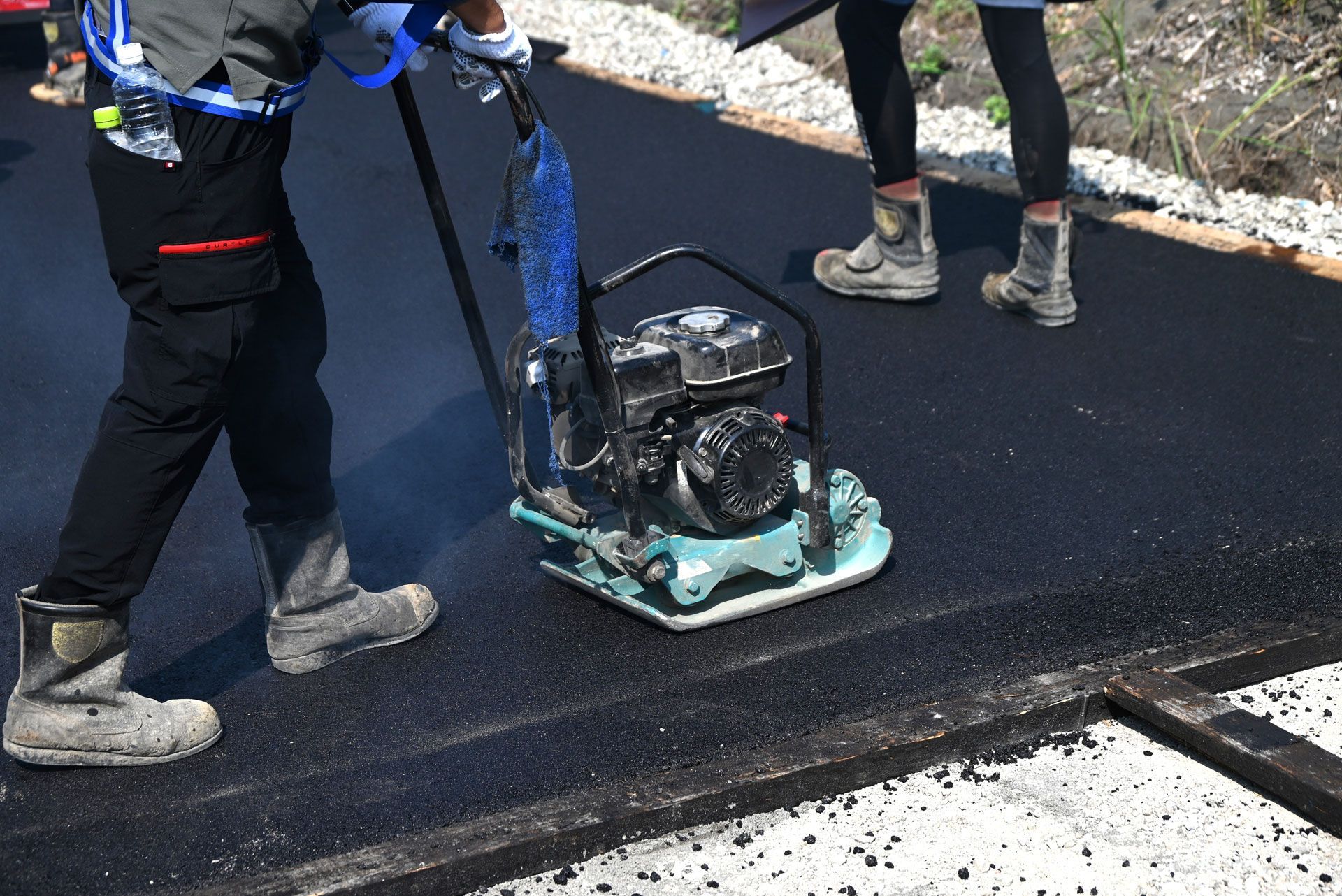 Worker using a handheld compactor on fresh black asphalt at a road construction site