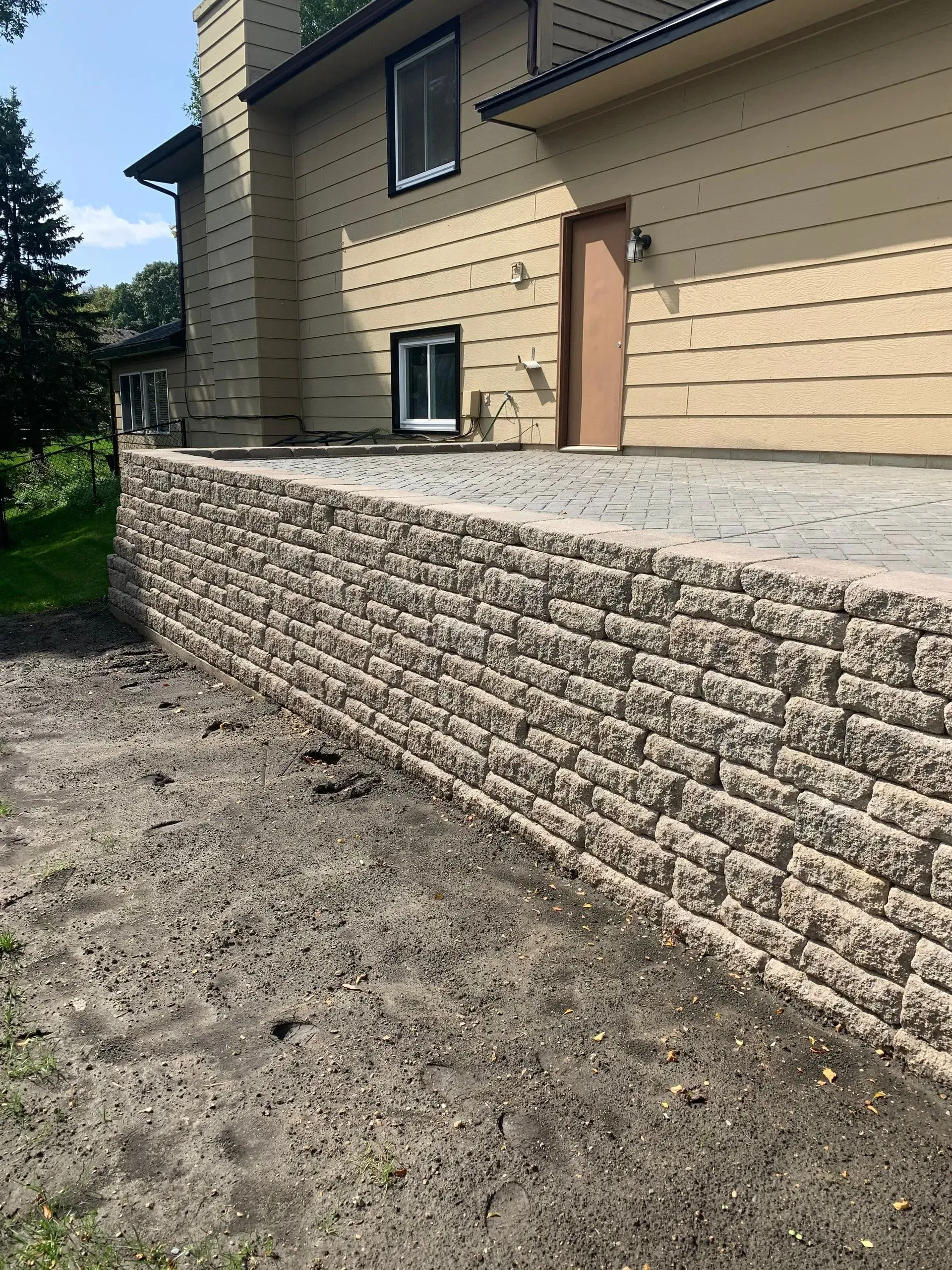 Beige block retaining wall along a building. Grey dirt area in front.
