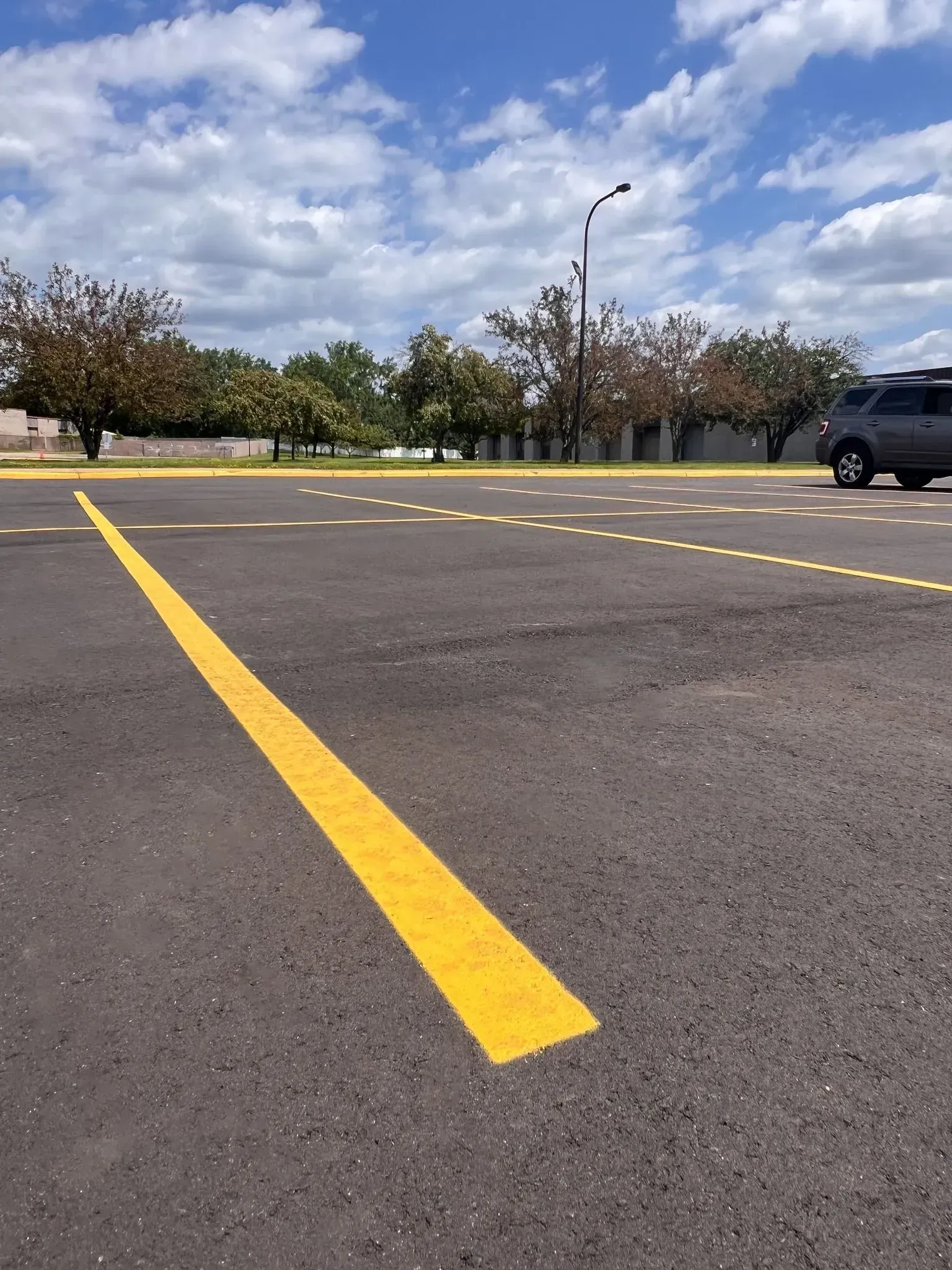 Yellow line on asphalt, indicating parking space in outdoor lot with car, trees, and cloudy sky.