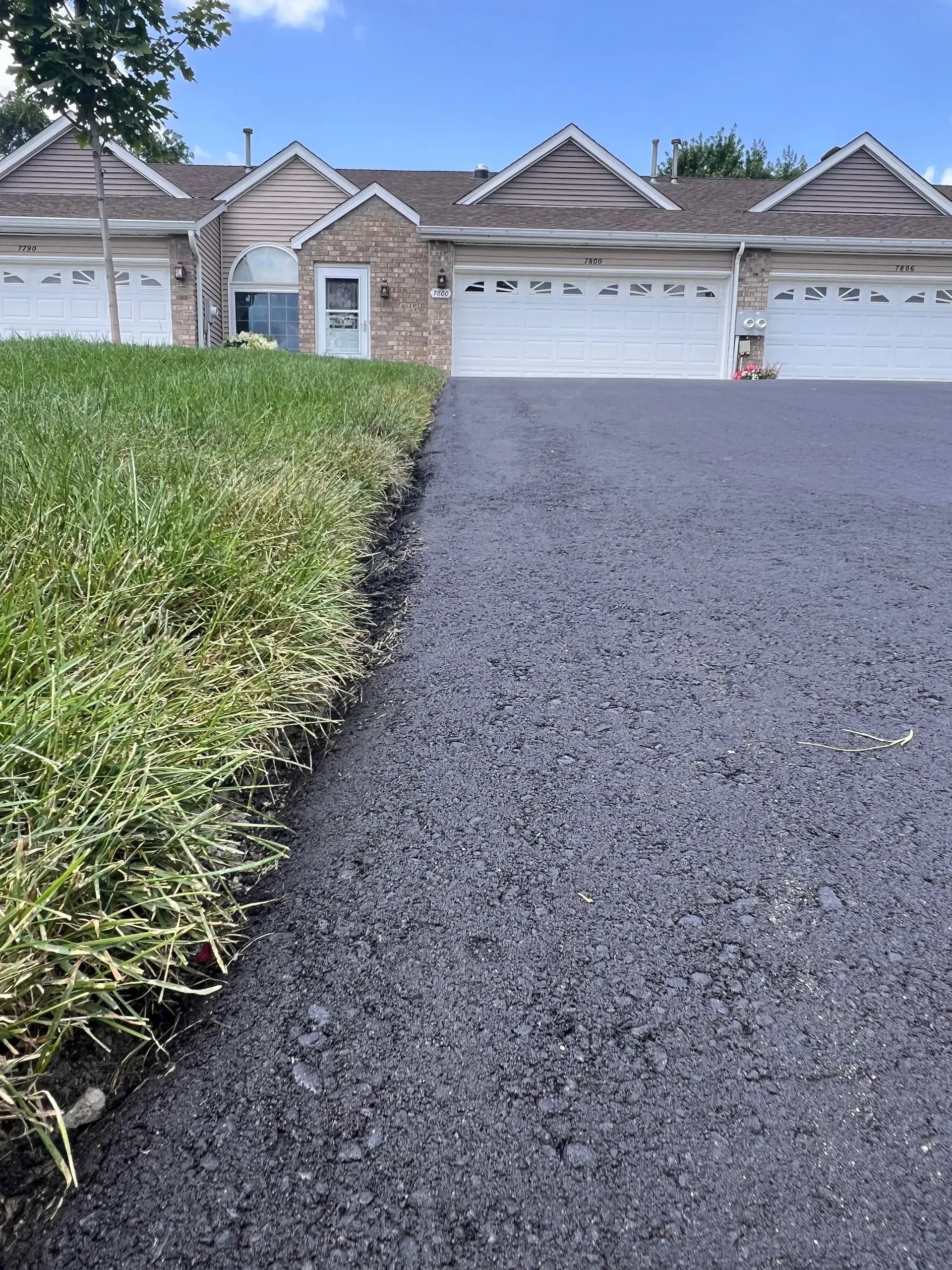 Asphalt driveway borders overgrown grass in front of townhouses with white garage doors.