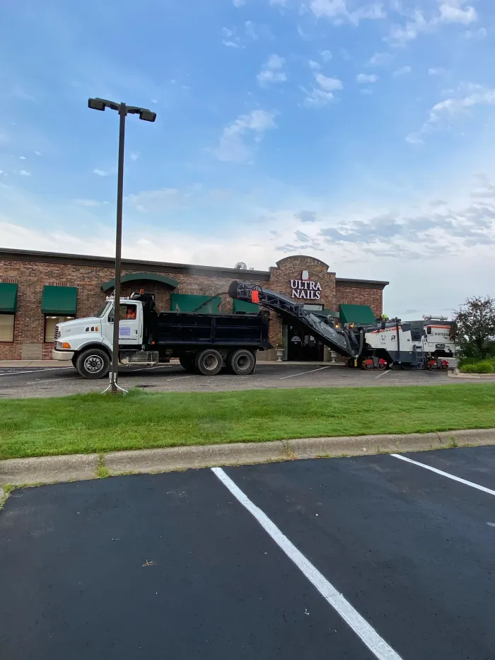 A dump truck being loaded with asphalt debris by a pavement milling machine in a parking lot.