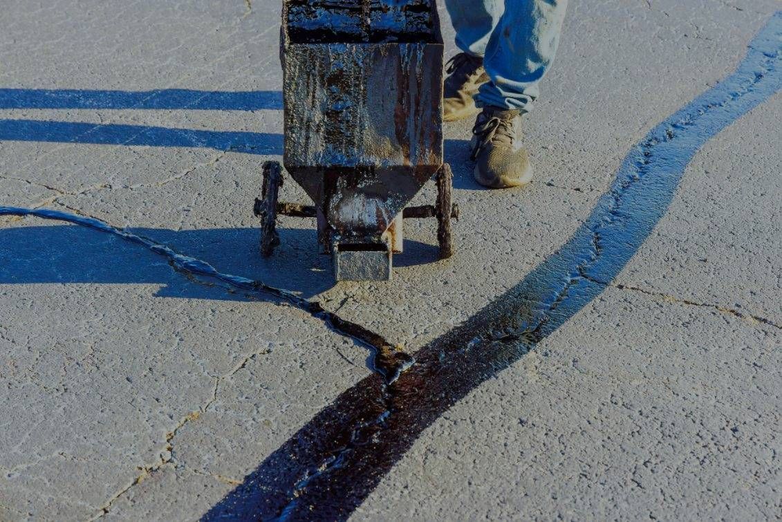 A person fills a crack in asphalt with a machine, leaving a dark, wet sealant.