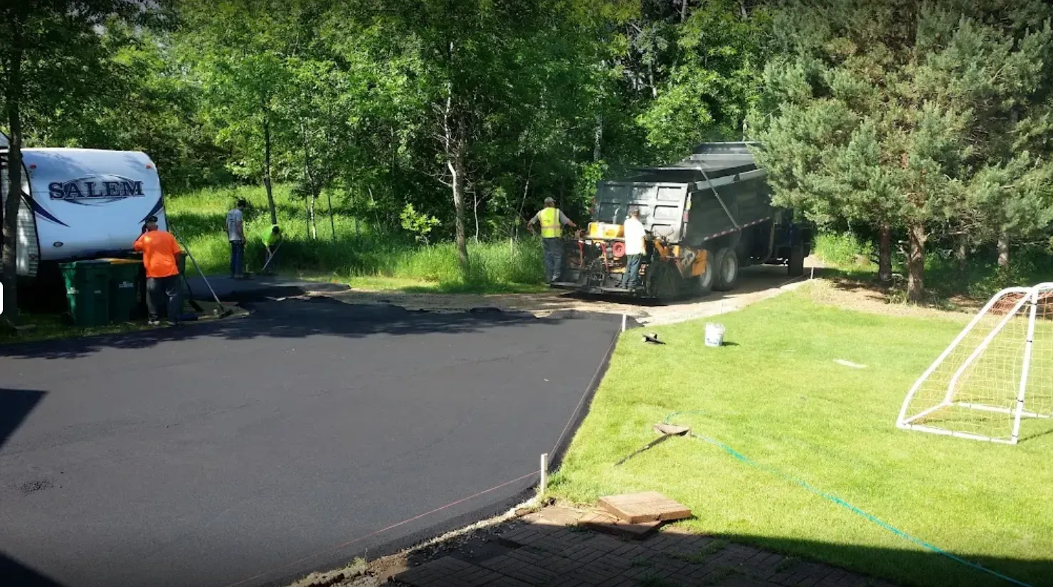Driveway paving in progress. Workers near trailer, fresh asphalt. Sunny day, green grass, trailer and trees in background.