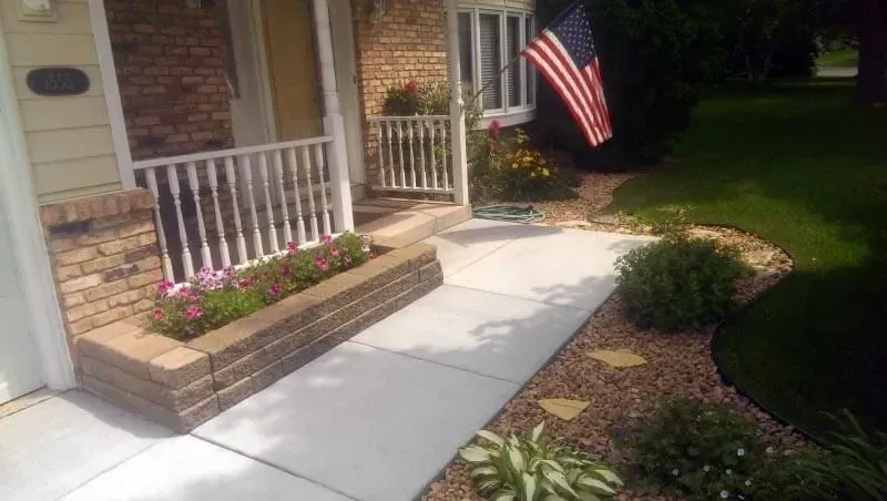 A brick house front with a concrete walkway, flower bed, and American flag.