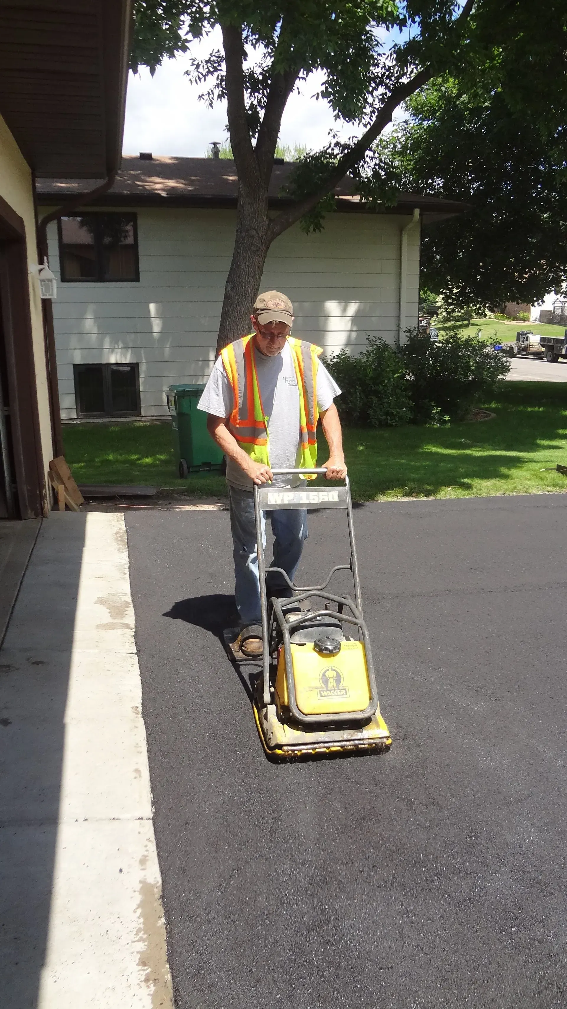 Man in orange vest compacting asphalt with a vibratory plate compactor on a driveway.