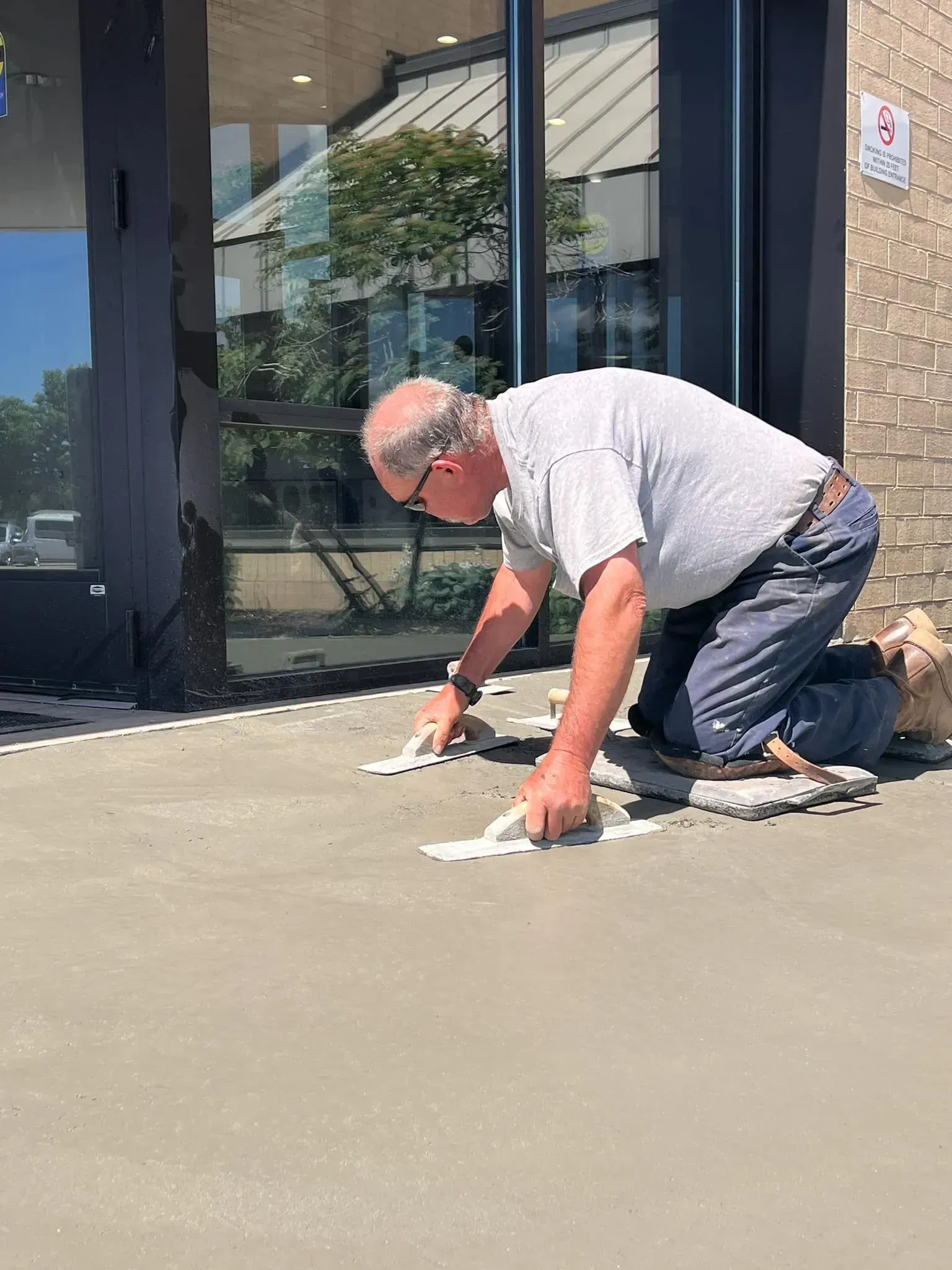 Man on knees placing tiles on concrete walkway, near a building.
