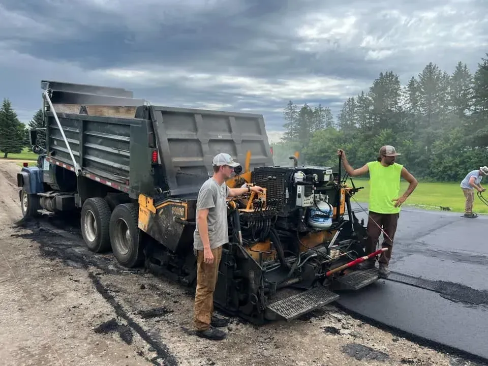 Workers paving a road with an asphalt paver machine, truck nearby, on a cloudy day.
