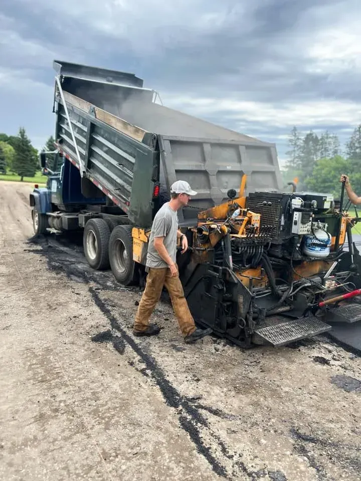 A dump truck dumping asphalt into a paving machine on a road; a man walks nearby.