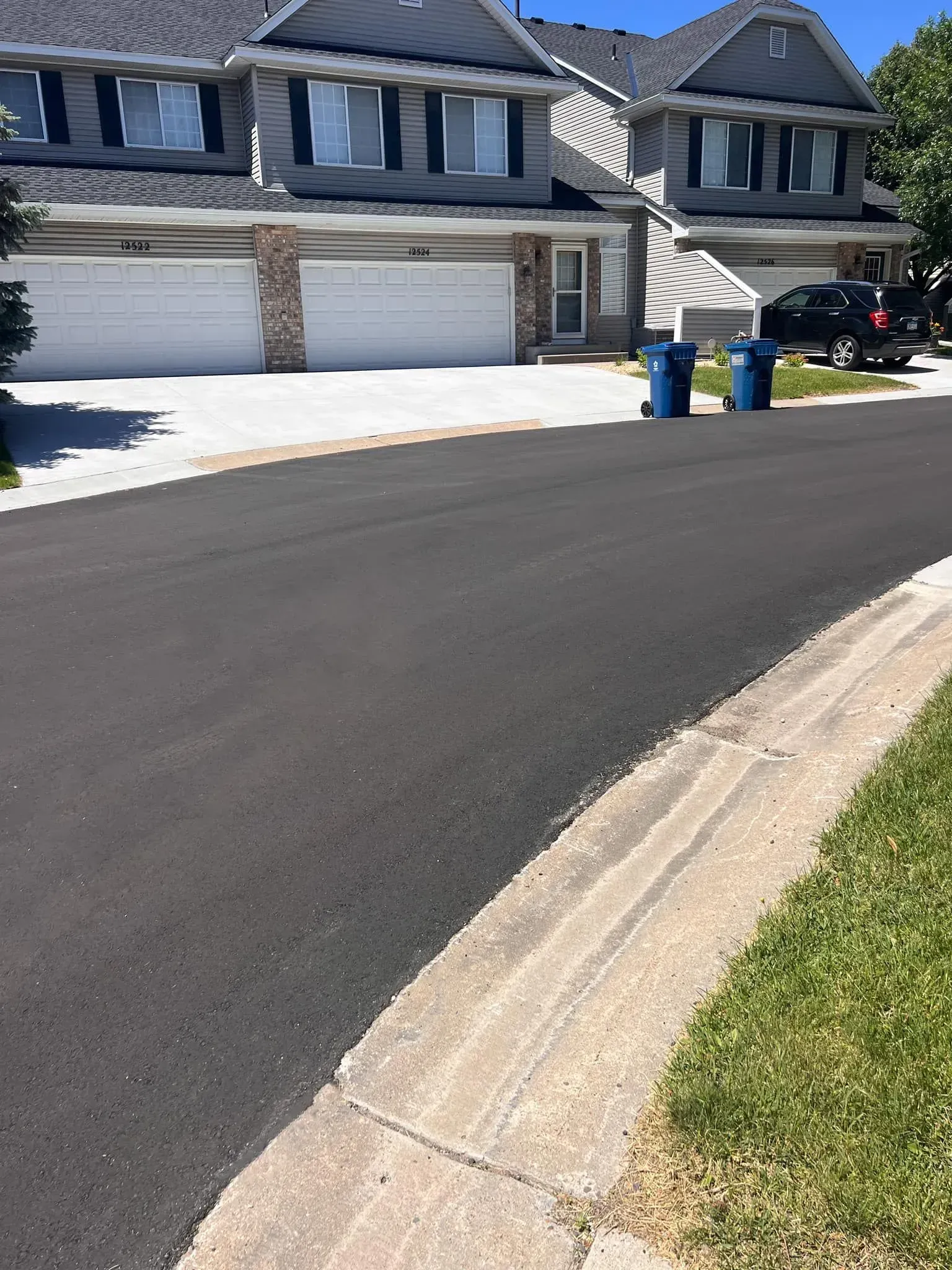 Asphalt road in front of townhomes with garages, blue trash cans, and a curb with grass.