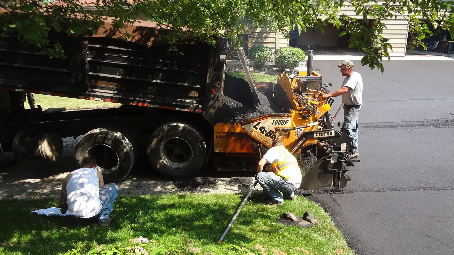 Workers paving a road: a truck dumps asphalt into a paving machine; two workers tend the machine, one kneels.