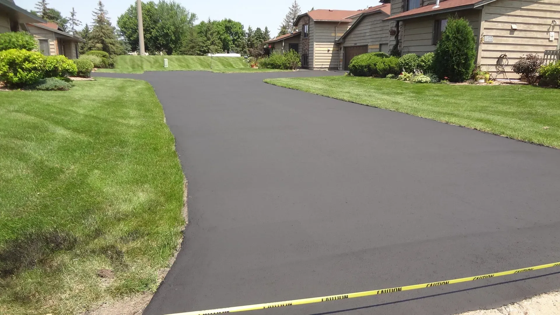 A newly paved black asphalt driveway in a residential neighborhood with green lawns and houses.