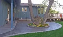 A patio with a tree in the center surrounded by flowers. The house is blue with a covered porch.