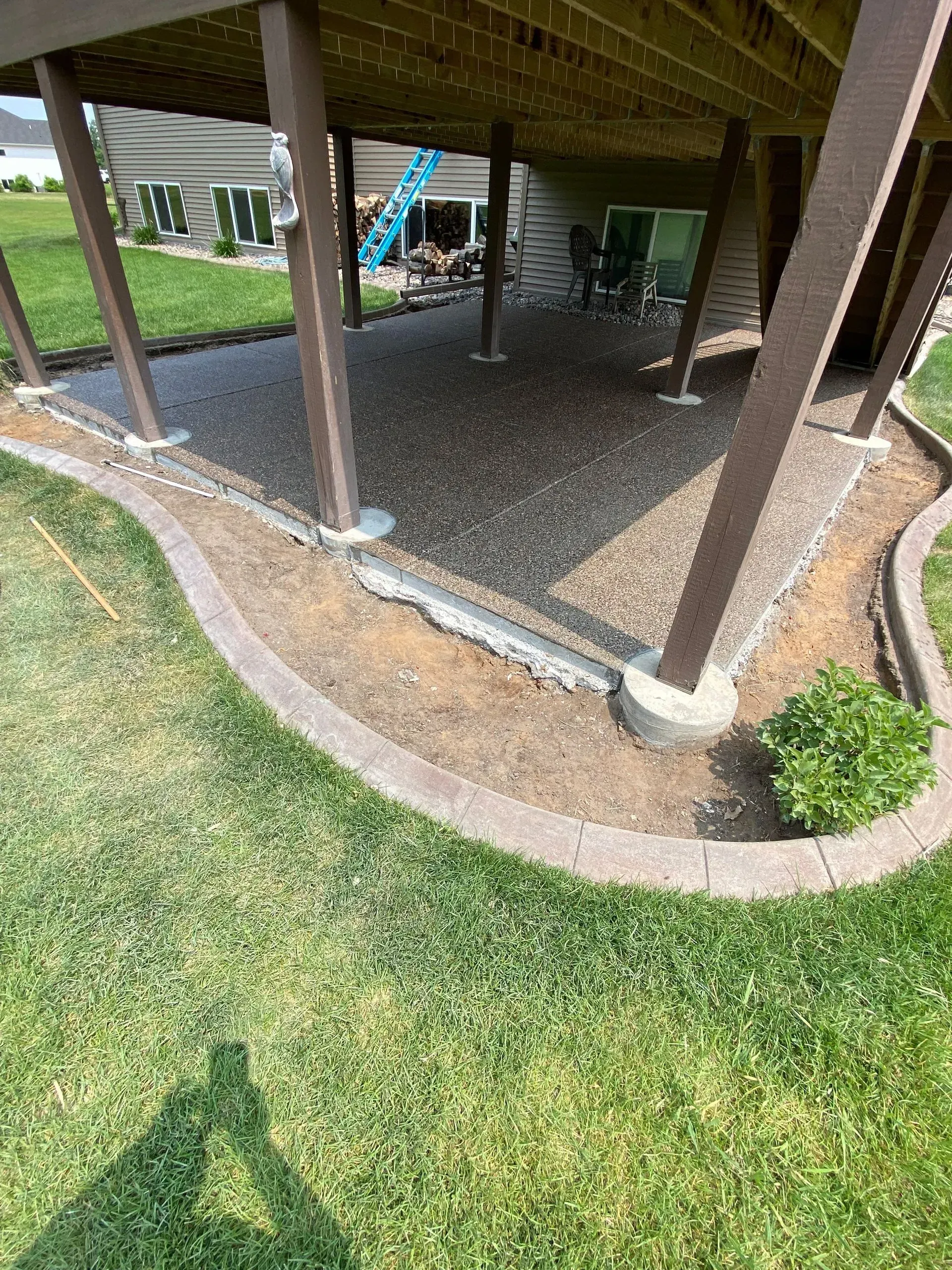 A gravel patio under a deck with brown columns, surrounded by a curved brick border and green lawn.