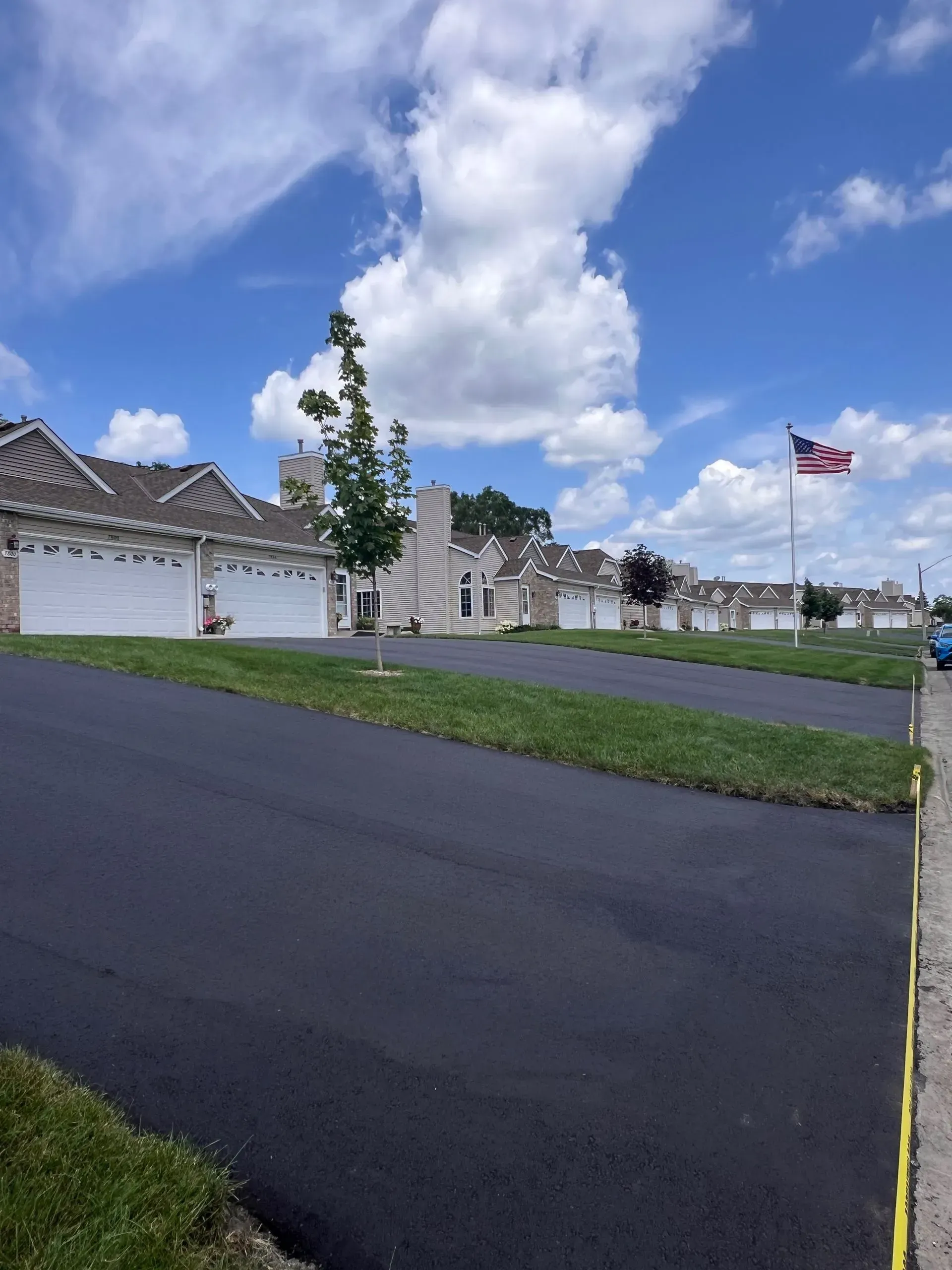 Row of houses with asphalt road, green grass, and American flag under blue sky.