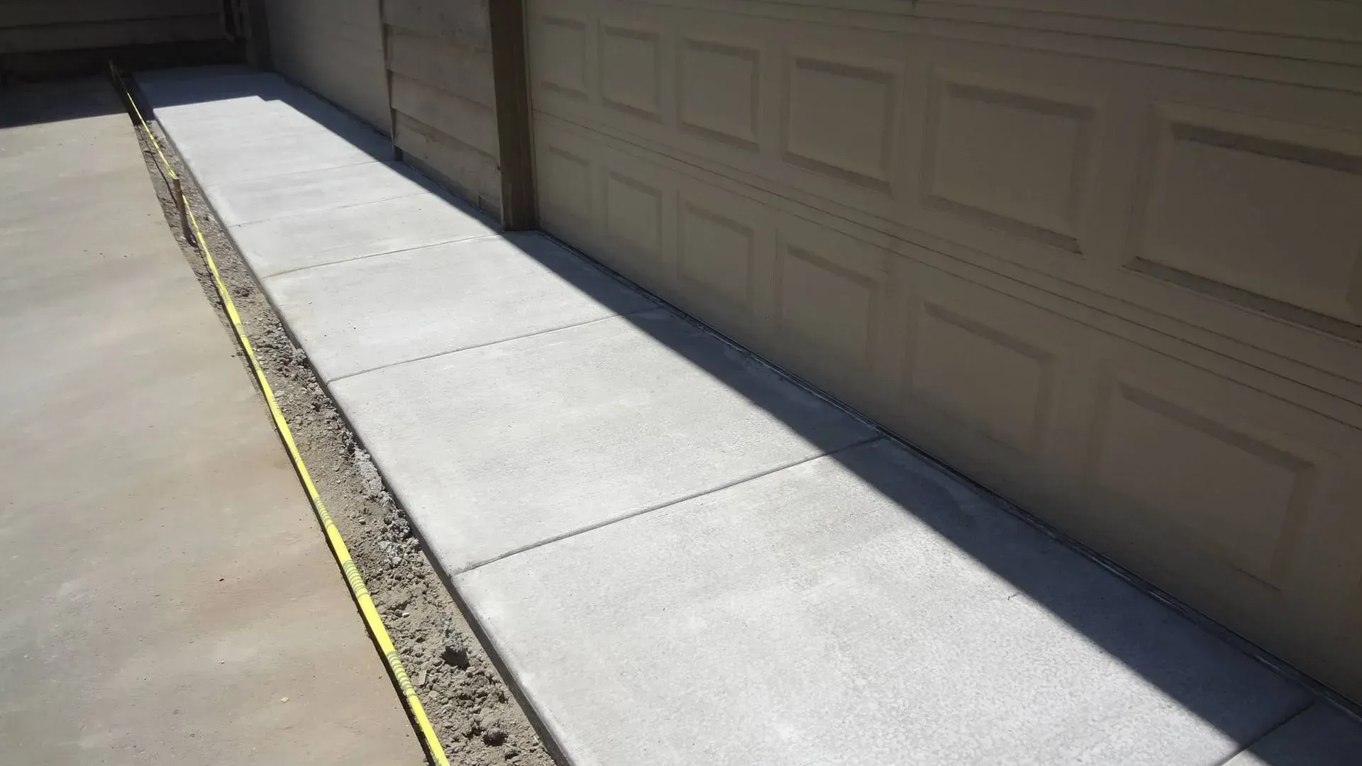 Concrete sidewalk next to a tan garage and wall, with a yellow line and dirt.