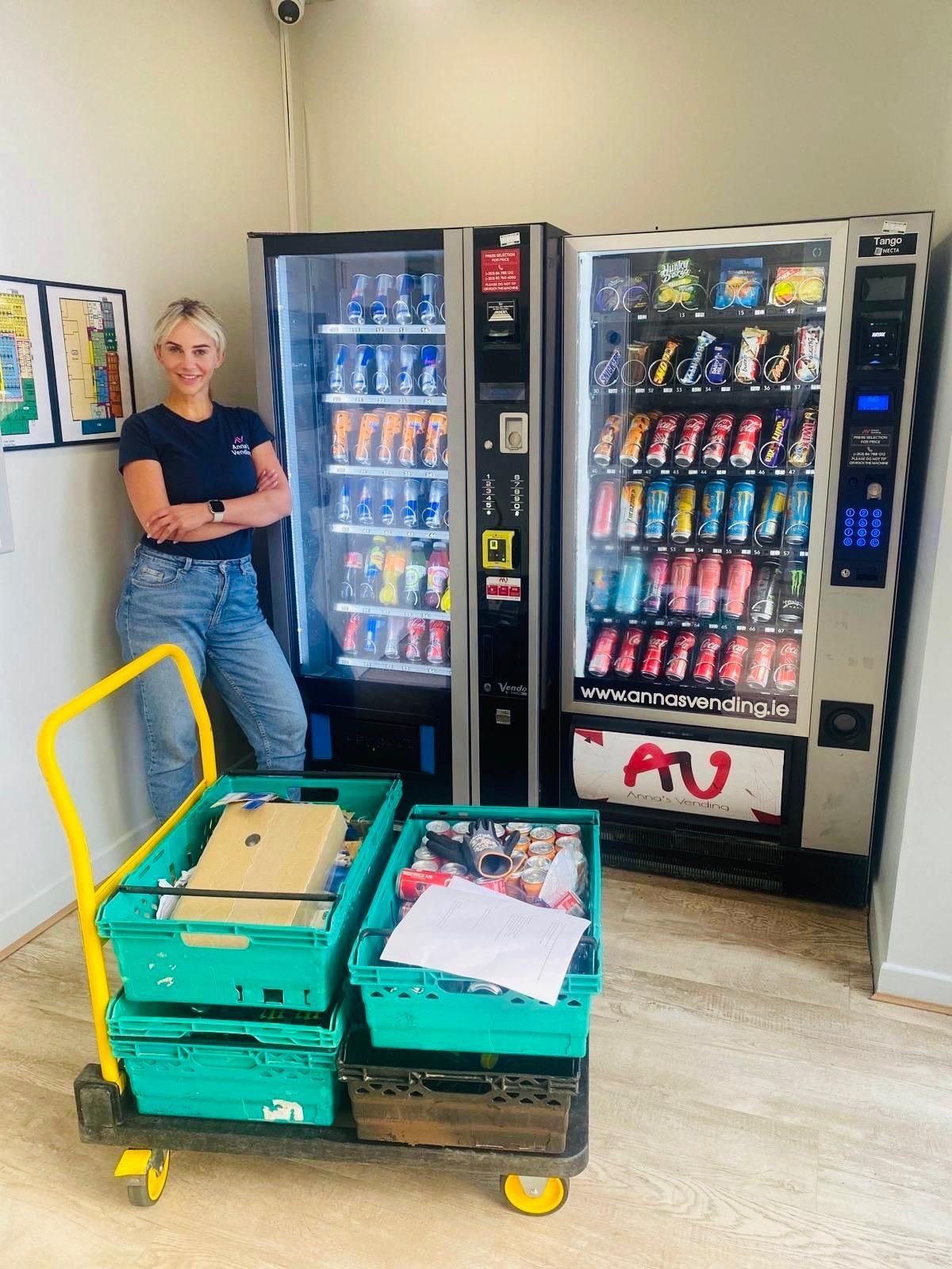 Woman standing beside snack and drink vending machines in a break room, with a cart of supplies in front.