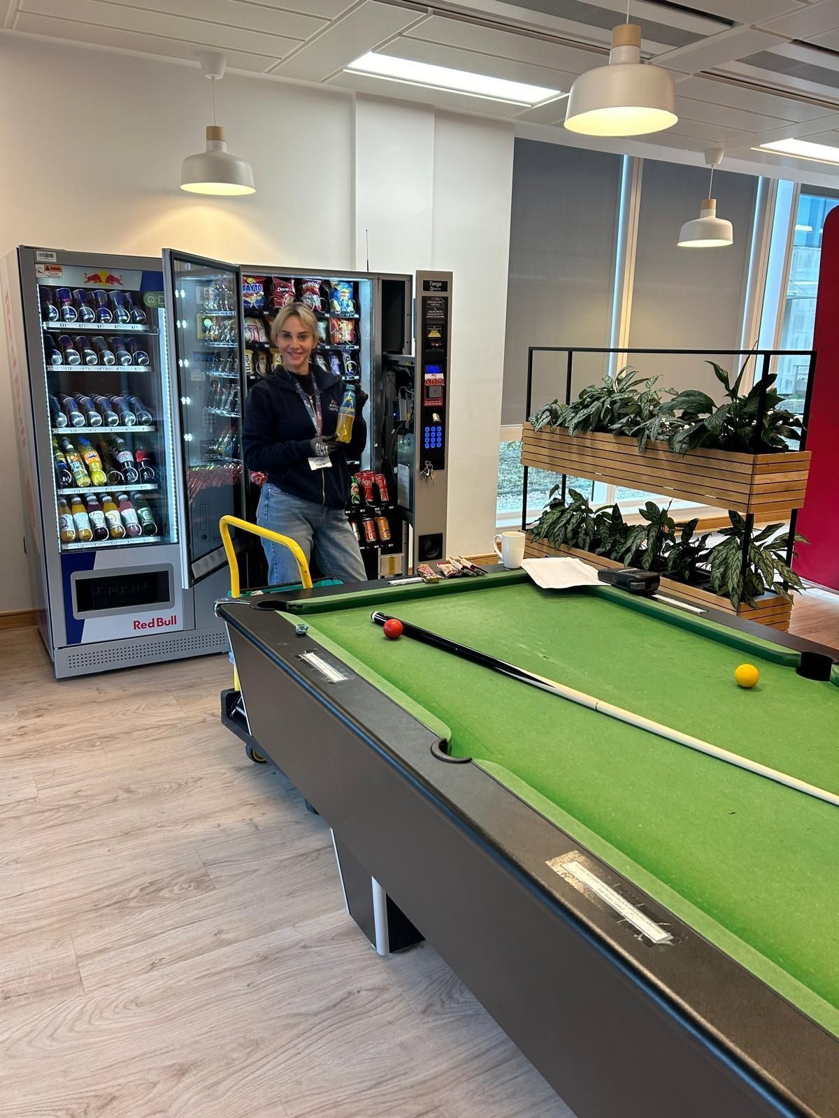 Man in an office lounge beside a pool table, with vending machines and hanging lights in the background