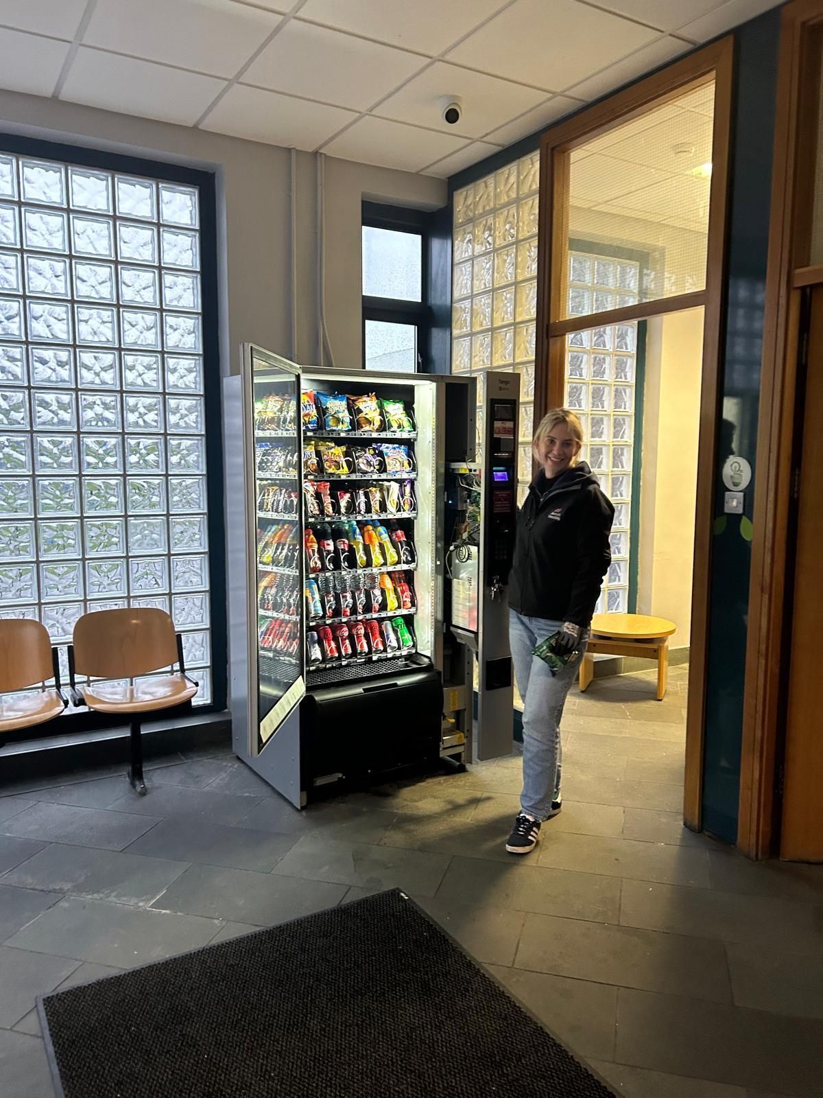 Person standing by a vending machine in a lobby with glass block windows and chairs.