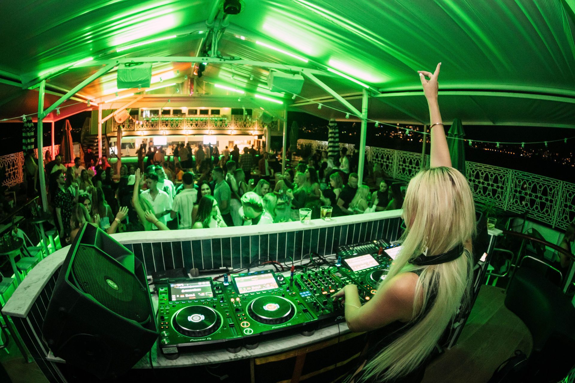 Emerald-lit rooftop crowd dances as DJ raises arm, view from behind the booth.