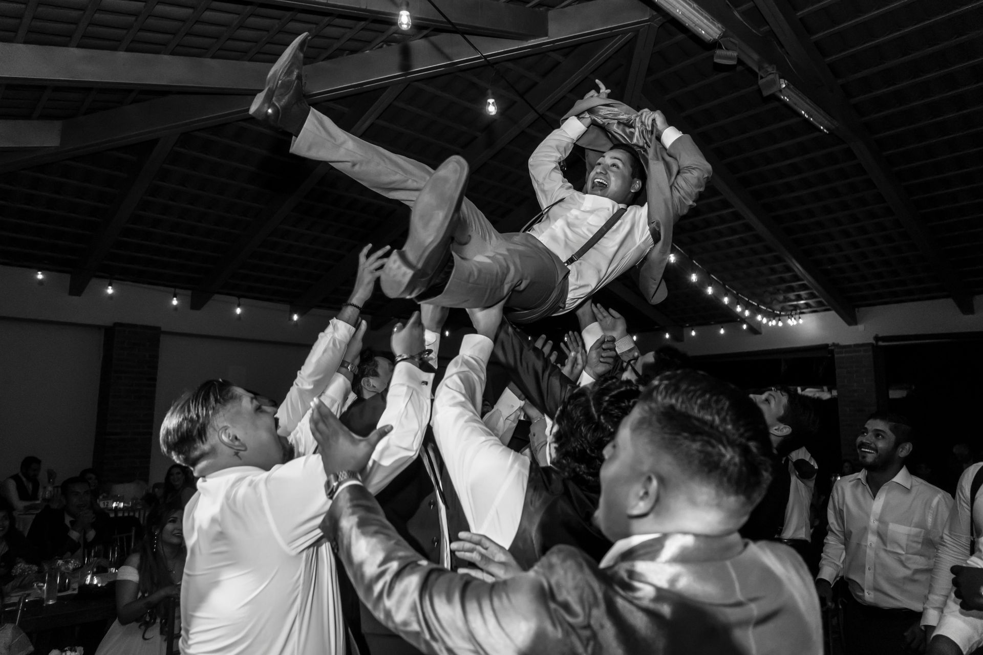 Groom being lifted and celebrated by groomsmen in suits at wedding reception with string lights.