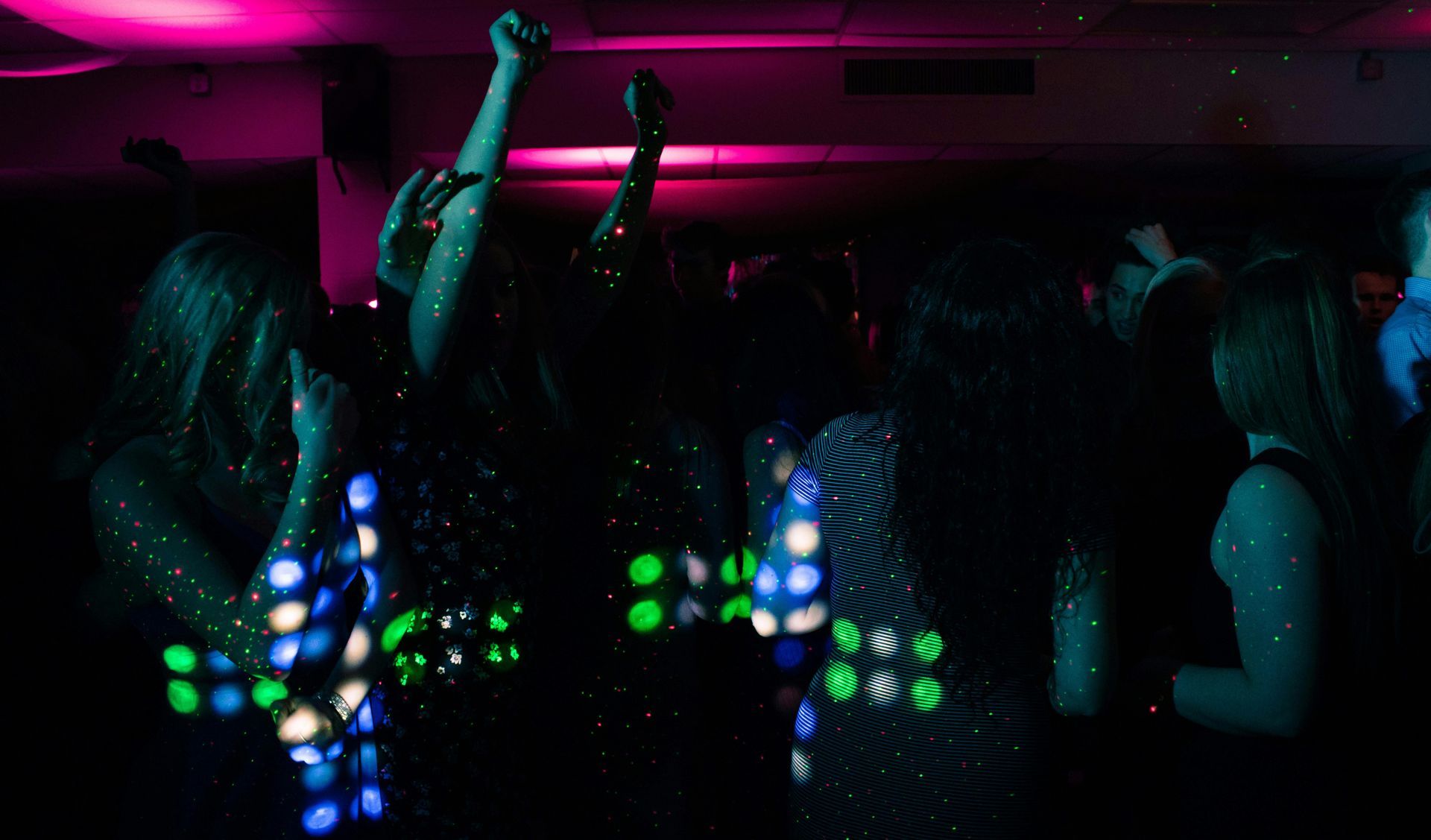 Silhouetted partygoers with arms raised dance under pink wash and green laser dots in packed club.