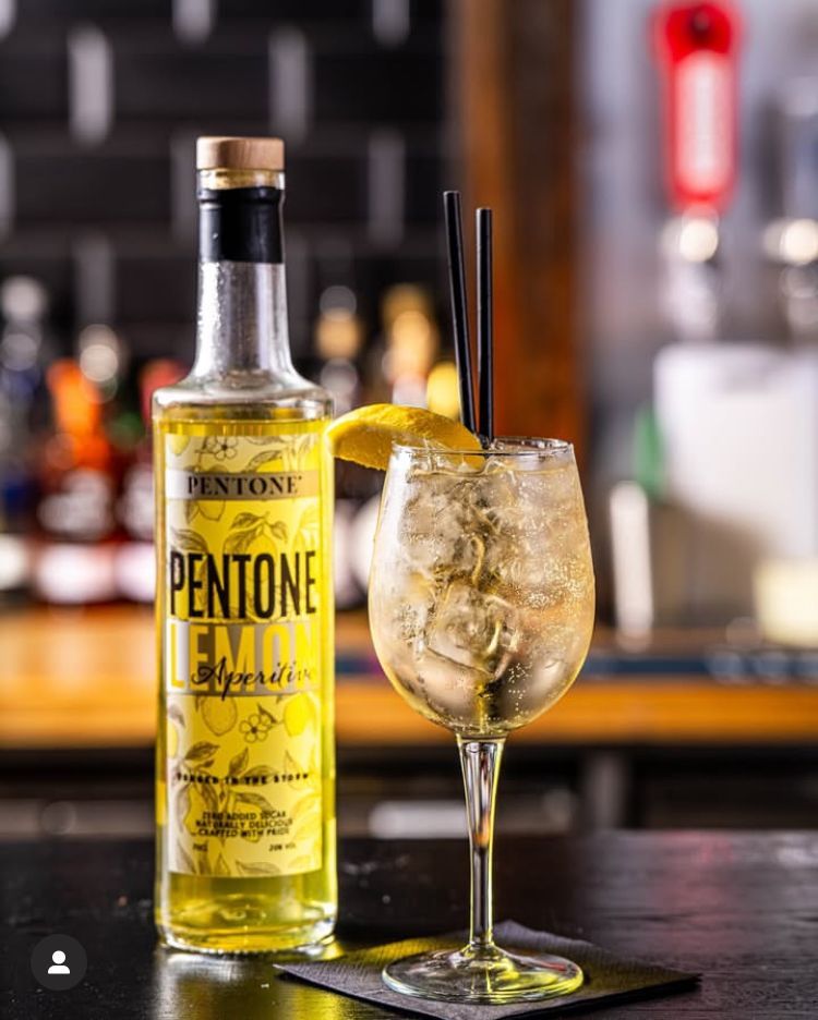 A bottle of Aperitvo Pentone Lemon, alongside a spritz glass with a double Pentone Lemon over ice, topped up with prosecco and soda, garnished with a slice of lemon.