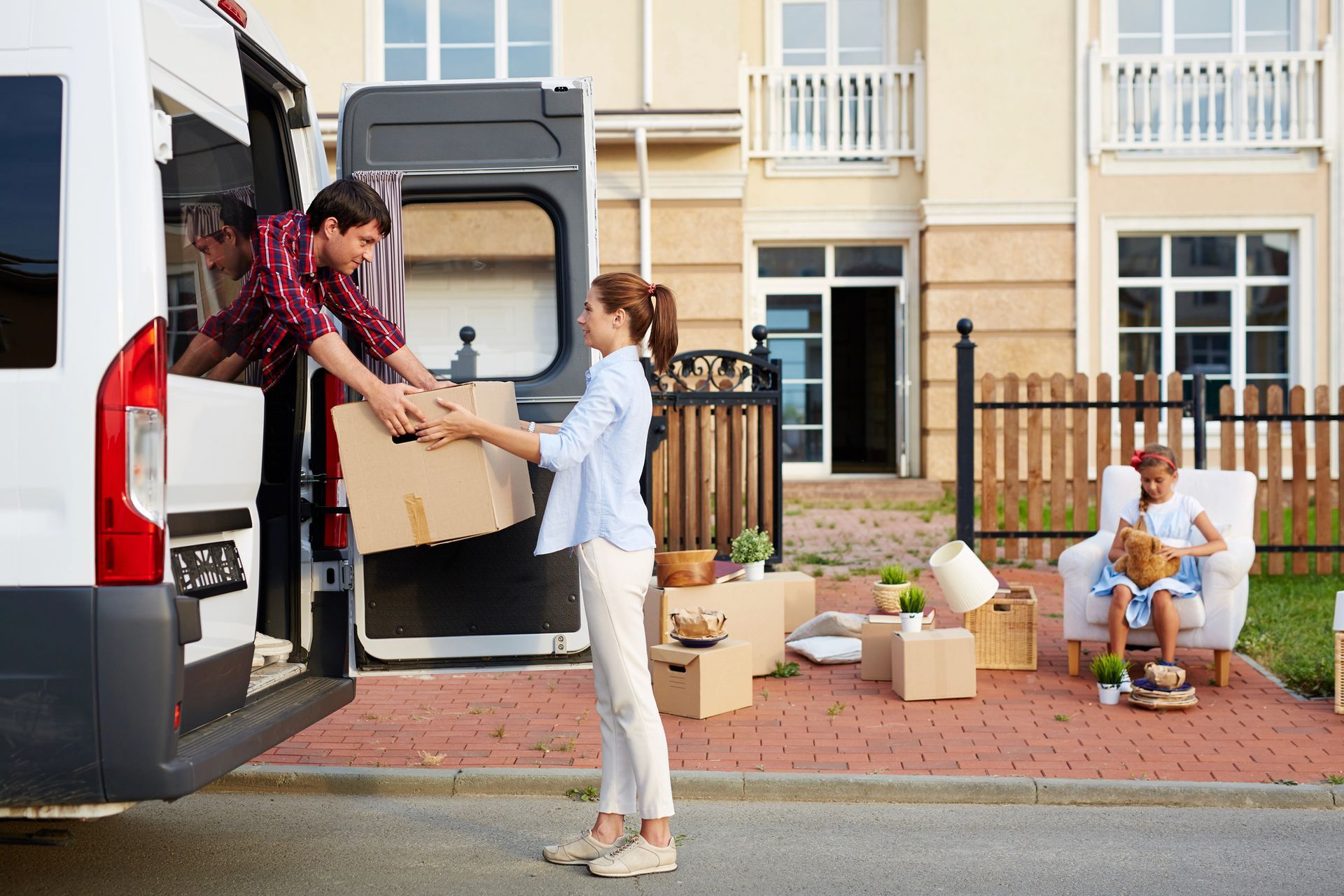 Persone che spostano scatoloni da un furgone a una nuova casa. Una donna riceve lo scatolone, altri lo caricano. Oggetti sparsi sul marciapiede.