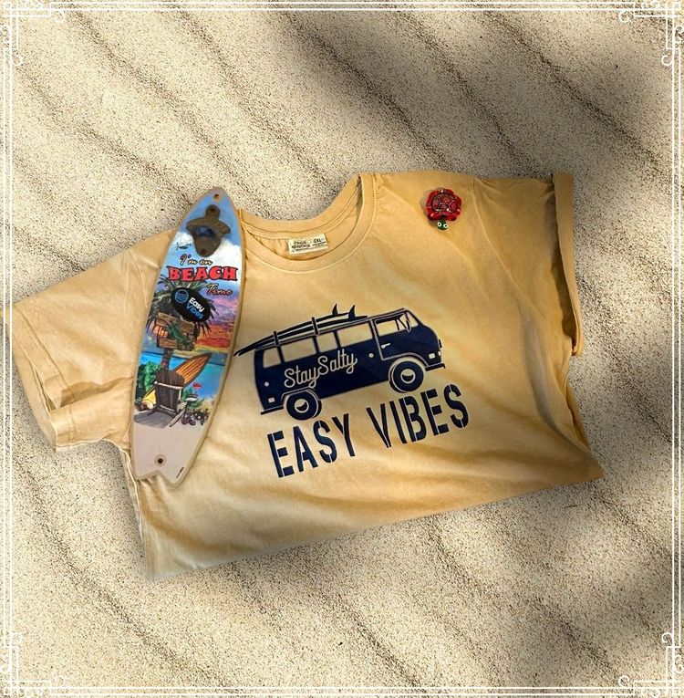 A beige Easy Vibes shirt featuring a VW Van in black printed on the front laying on a sandy beach, shown with a mini surfboard laying on it 