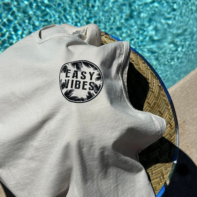 Black Easy Vibes logo on beige shirt laying on a straw hat beside a pool