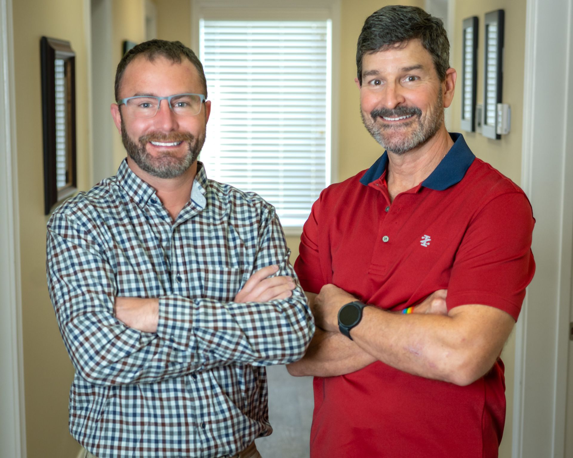 Two men are standing next to each other in a hallway with their arms crossed.