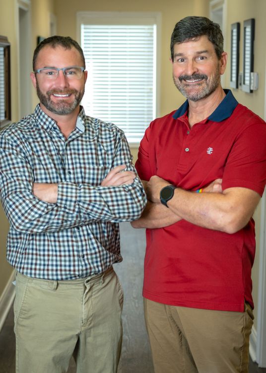 Two men are standing next to each other in a hallway with their arms crossed.