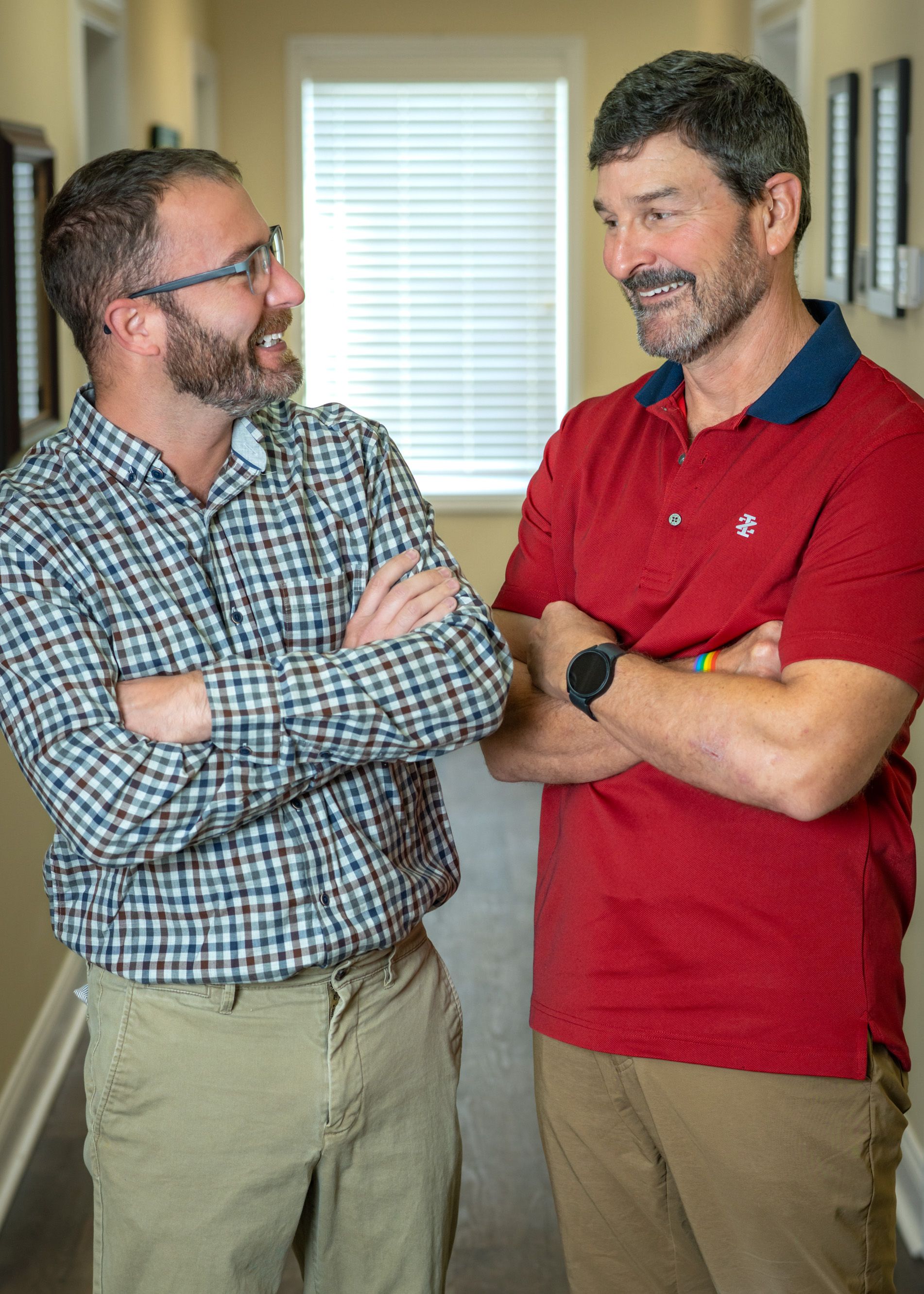 Two men are standing next to each other in a hallway with their arms crossed.