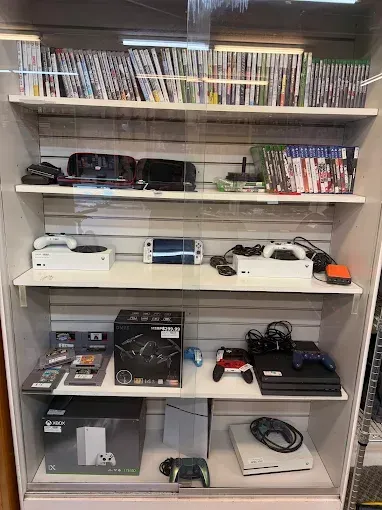 A display case filled with video game consoles and games. Consoles are black and white, games lined on shelves.