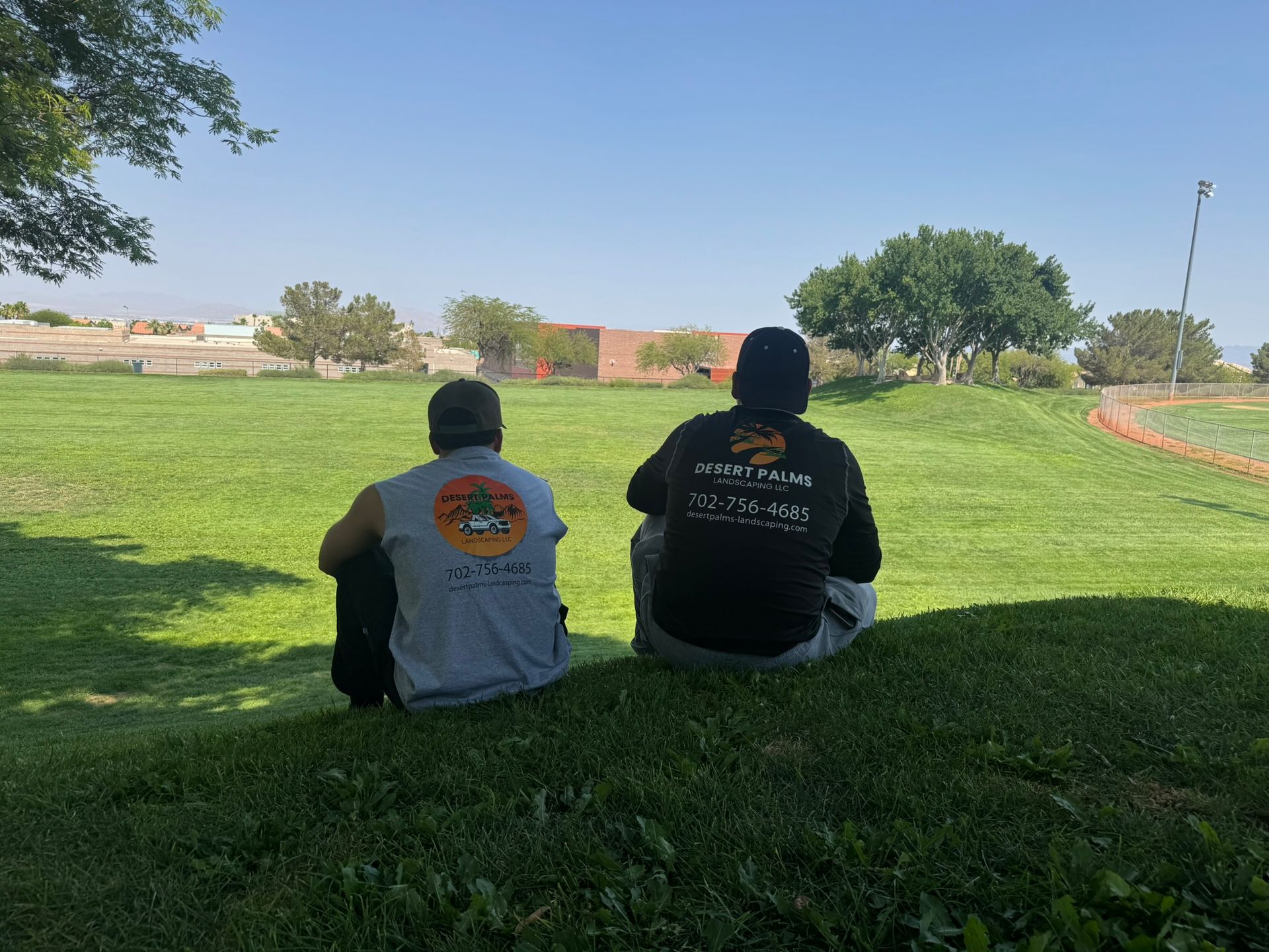 Two landscapers sit on a grassy hill, backs to the camera, facing a green field under a blue sky. They appear to be taking a break.