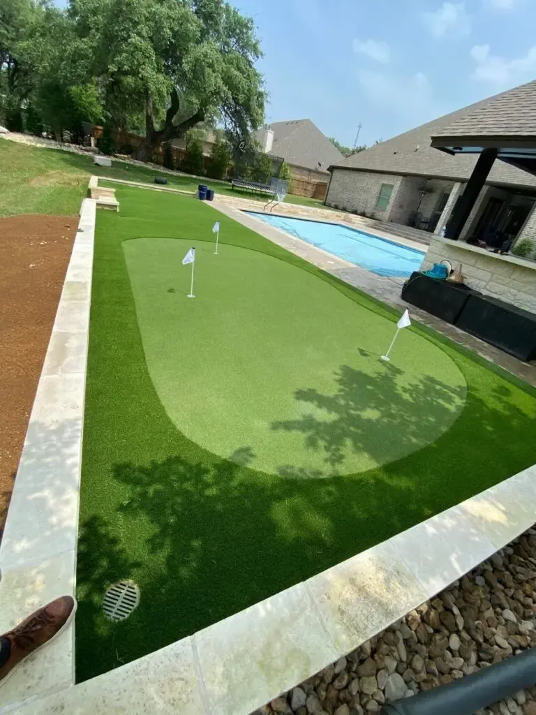 A backyard putting green with white flags, next to a pool and a house on a sunny day.