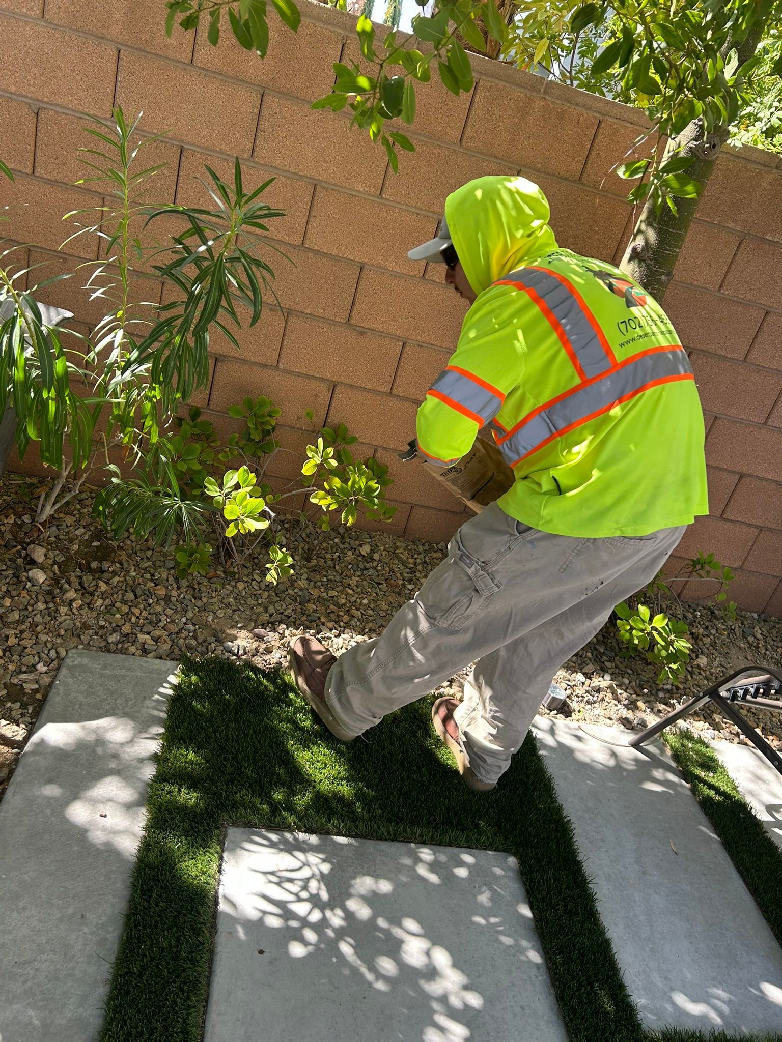 Person in neon safety vest and hat bends down near stone pavers and greenery, in front of a brown brick wall.