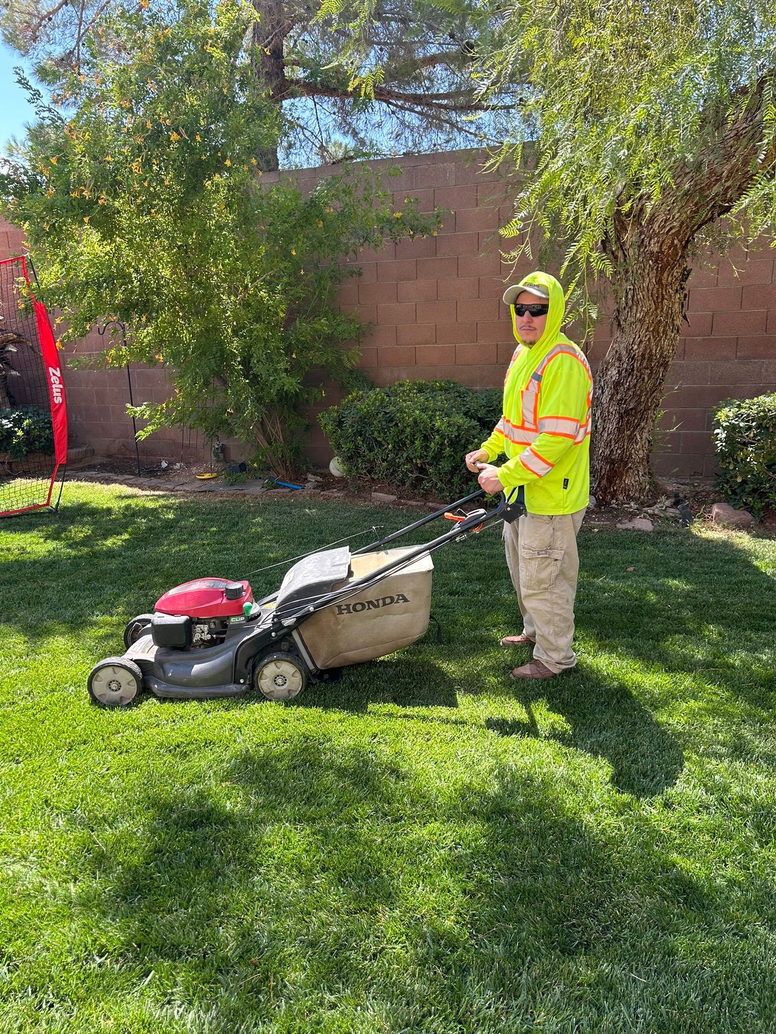 Person mowing a lawn in a yard; wearing a yellow reflective jacket on a sunny day.