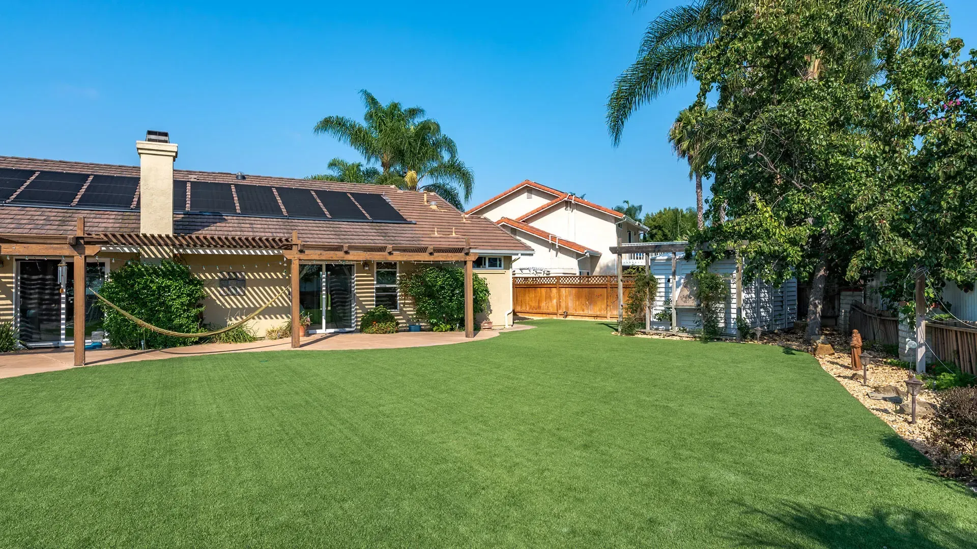 Backyard with green lawn, patio, house with solar panels, and trees on a sunny day.