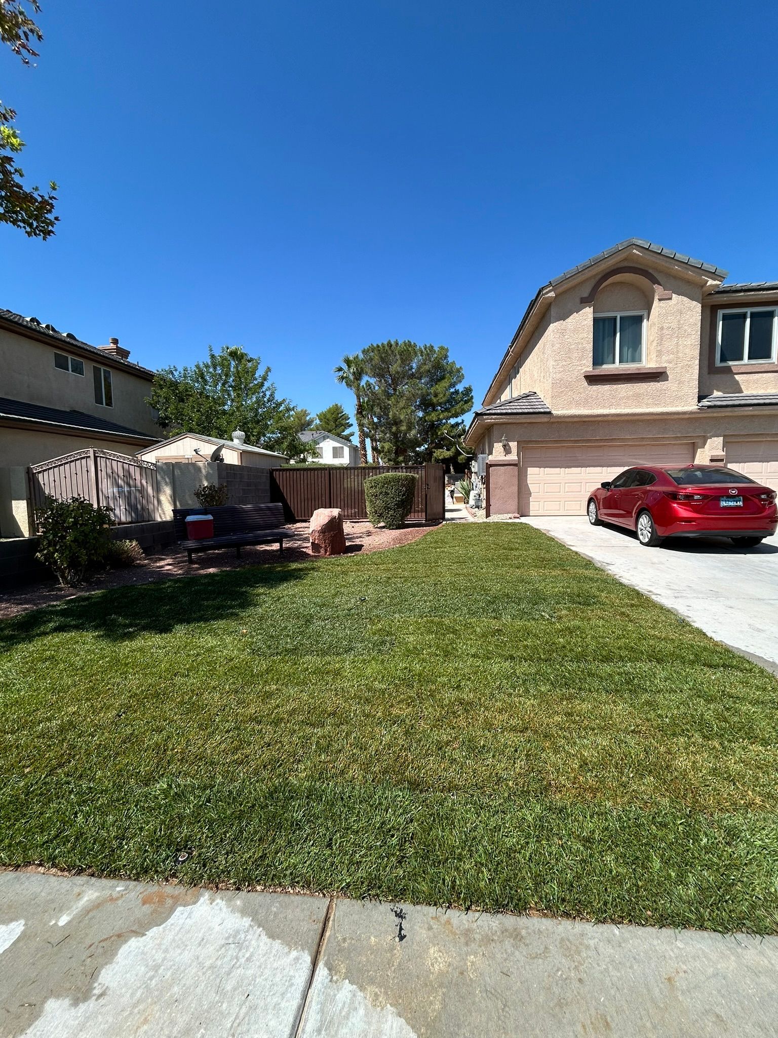 Green lawn in front of beige two-story house with a red car parked in the driveway under a clear blue sky.