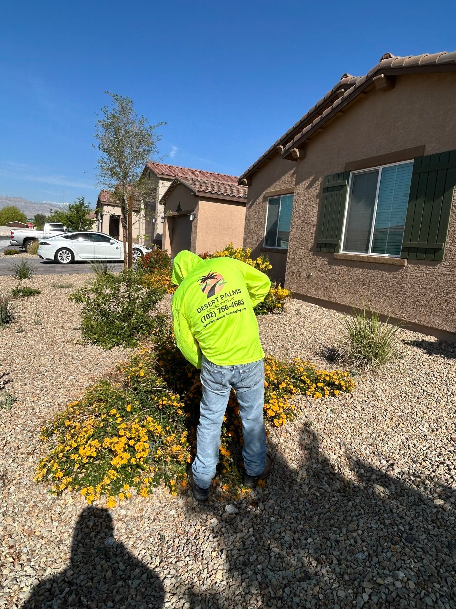 Person in a neon green hoodie tending a flower bed near a house on a sunny day.