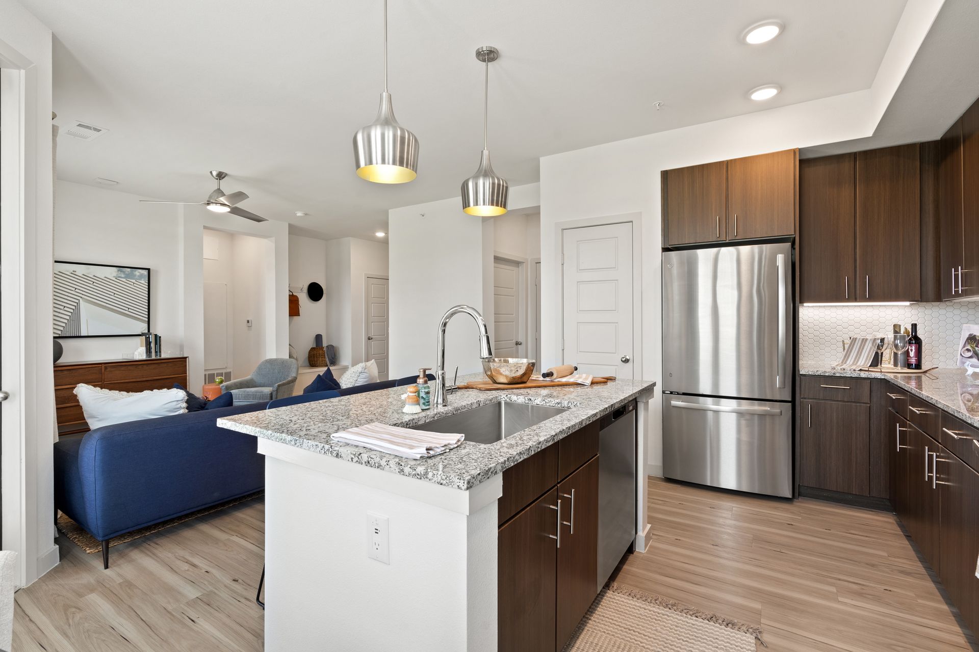 Open-concept kitchen with granite island, stainless steel fridge, and living area in background.