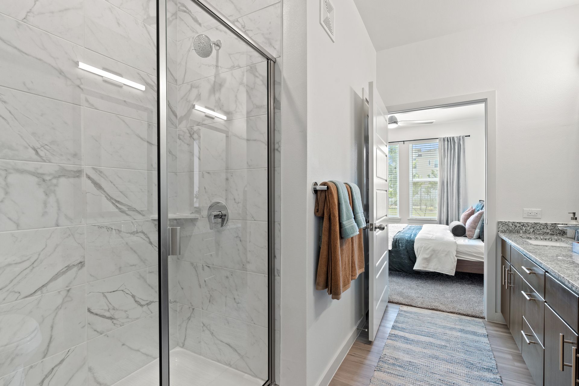 Glass-enclosed marble shower in a modern apartment bathroom with a view into the bedroom.