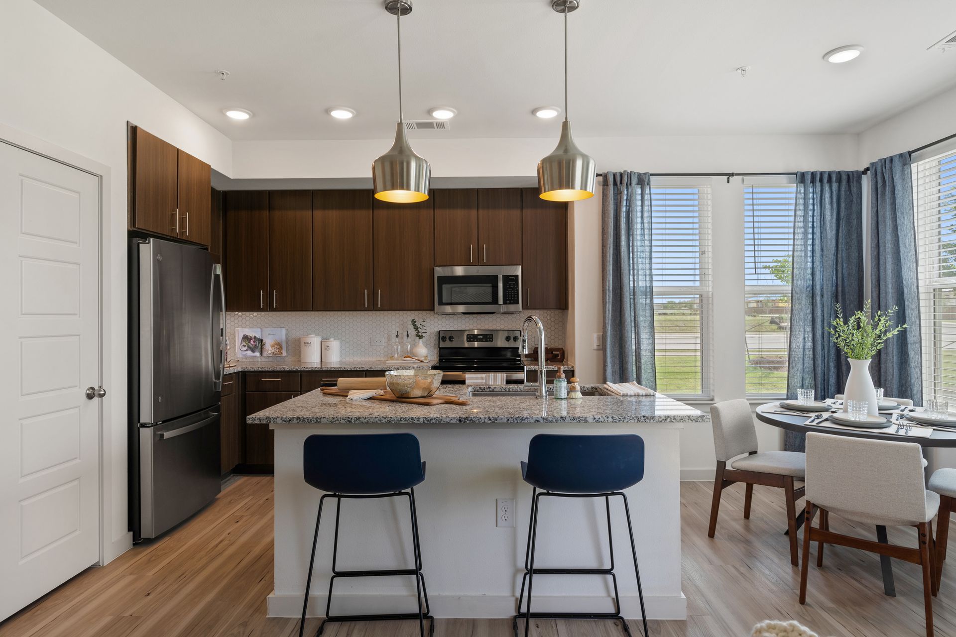 Open-concept kitchen with granite island, dark wood cabinets, stainless appliances, and blue bar stools.